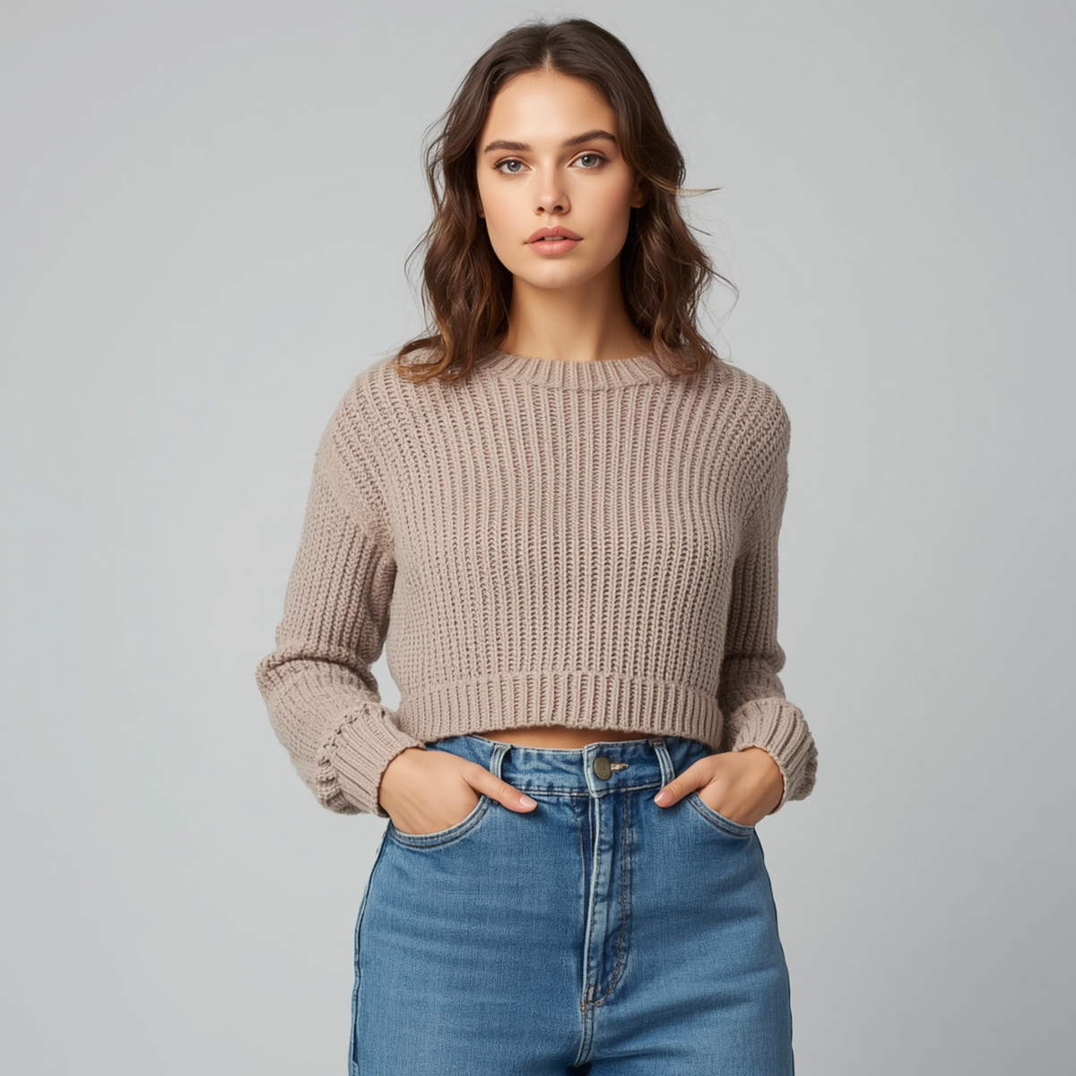 Frau trägt einen beigen Crop-Pullover mit Rippstrickstruktur, Rundhalsausschnitt und langen Ärmeln zu blauen Jeans vor hellem Hintergrund.